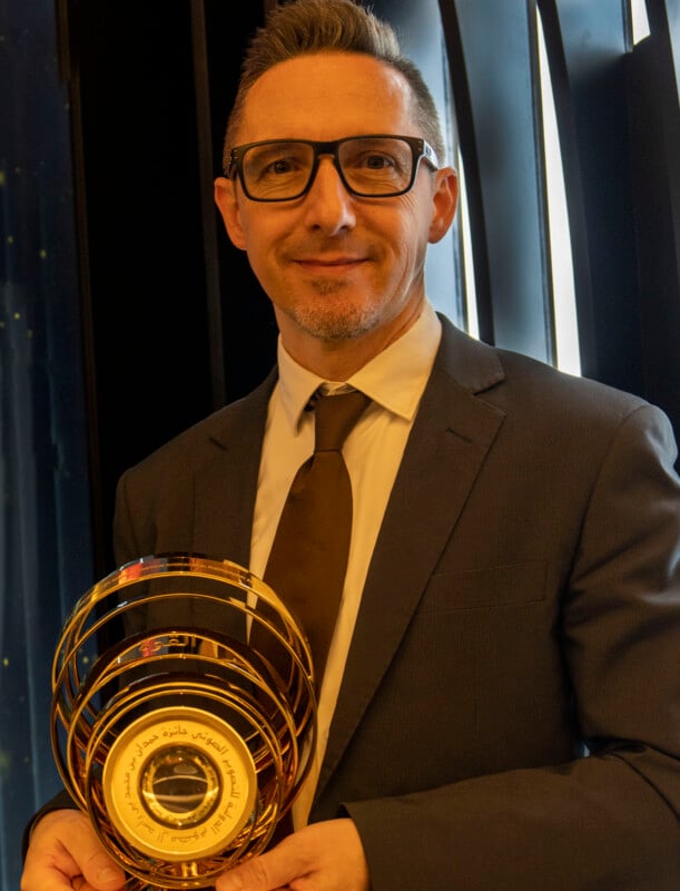 Un hombre sonriente vestido con traje, corbata y gafas sostiene una medalla de cristal dorada con escritura árabe. El fondo presenta paneles abstractos y modernos en negro y azul.