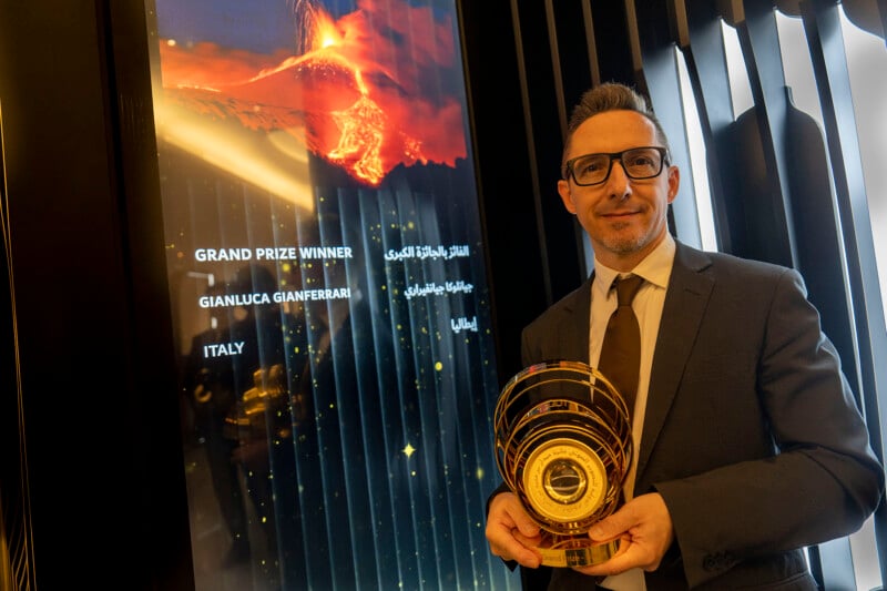 Un hombre con traje y gafas estaba de pie junto a una pantalla que sostenía un trofeo dorado en el que aparecía su nombre: Gianluca Gianferrari, el ganador del gran premio de Italia. El fondo muestra una vívida imagen de una erupción volcánica.