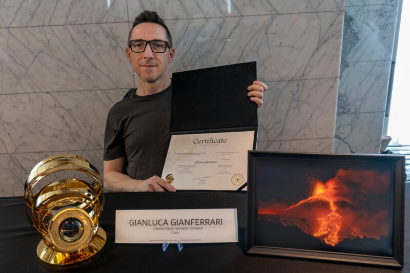 Un hombre con gafas está sentado a una mesa que muestra un certificado público, un trofeo de oro y una fotografía enmarcada de un volcán incandescente. La placa de identificación dice "Gianluca Gianferrari, ganador del Gran Premio del iPhone, Italia.