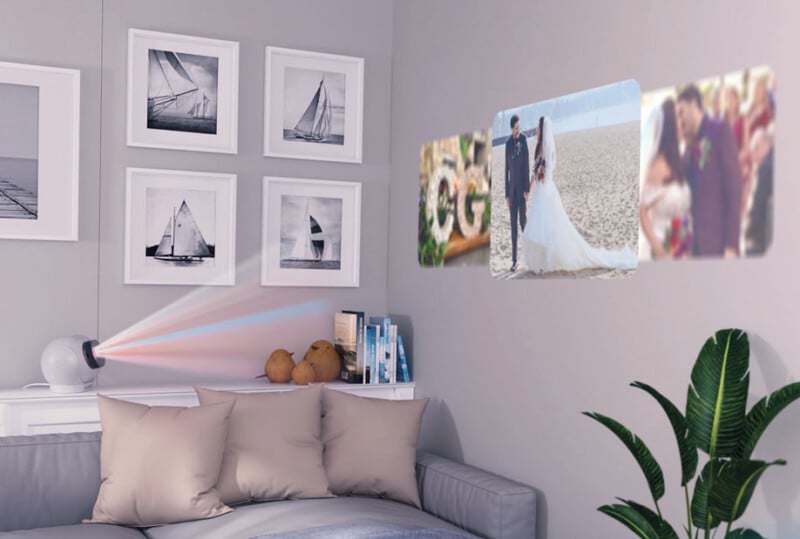 Hay un pequeño proyector en la pared del salón y un sofá con cojines beige que muestran fotos de boda. Cerca cuelga una imagen enmarcada de un velero y en una esquina hay una planta.