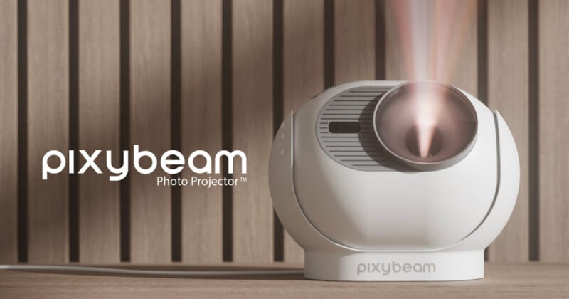 Un proyector esférico blanco etiquetado "haz de elfo" Emite un haz de luz brillante. El proyector se sitúa sobre una superficie de madera con un fondo de lamas verticales. texto "proyector de fotos pixybeam" aparece a la izquierda.