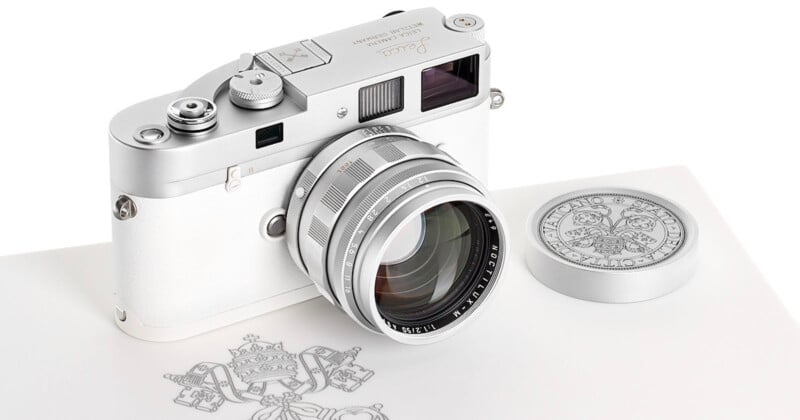 Una cámara Leica plateada y blanca con lente y cubierta a juego descansa sobre una superficie con emblemas ornamentados y líneas detalladas. La cámara presenta elementos de diseño clásicos y elegantes.