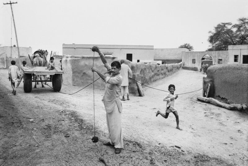A man spins a top with string while smiling at the camera in a rural village. A child runs nearby, and an ox cart with people is in the background along with mud-walled houses and trees.