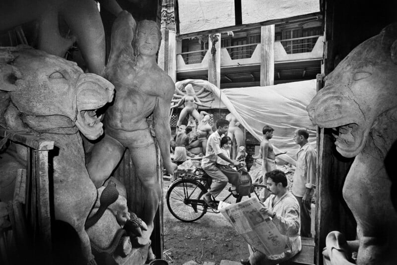 A busy street scene viewed from inside a sculptor’s workshop, with unfinished statues of lions and human figures in the foreground, people walking and a man riding a bicycle in the background, and a man reading a newspaper.