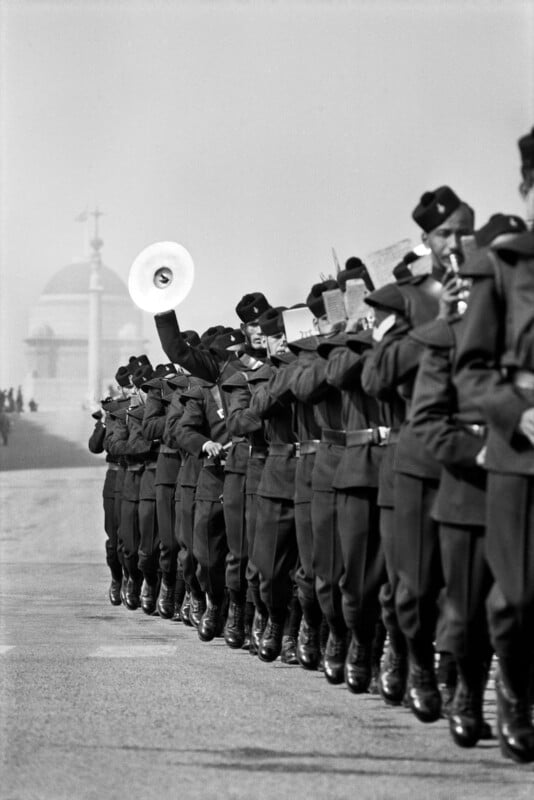 A black-and-white photo of a military band marching in formation outdoors; one member holds a cymbal aloft while others play instruments. A domed building is visible in the background.