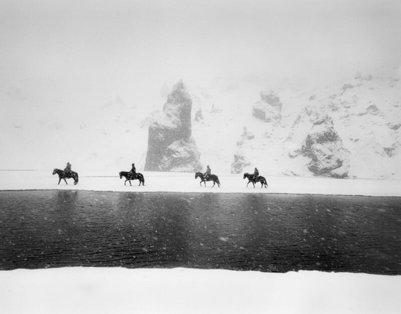 Four people on horseback ride along a snowy, windswept riverbank, with large rocky formations and snow-covered mountains shrouded in mist in the background.