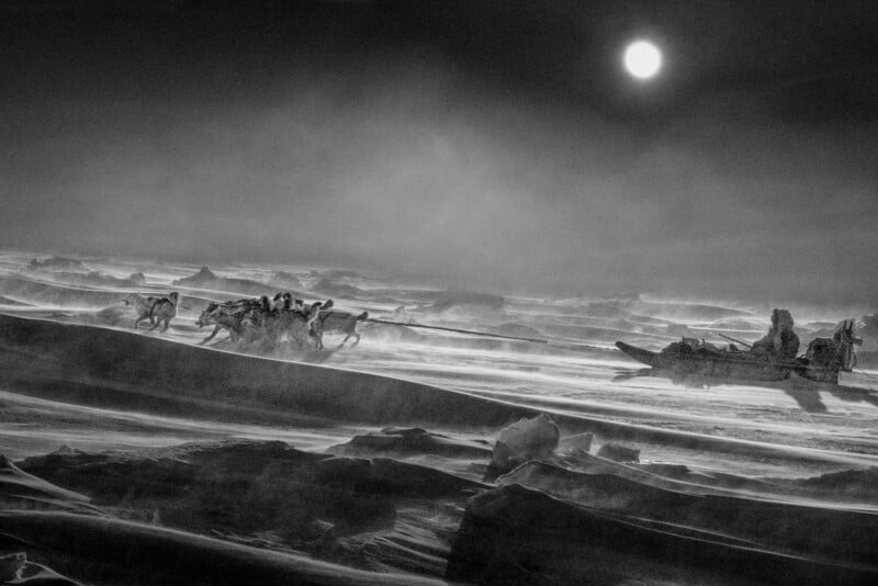 A black and white photo of a dog sled team racing across a snowy, windswept arctic landscape under a bright sun in a cloudy sky. Two people ride the sled, surrounded by dramatic drifts and icy terrain.