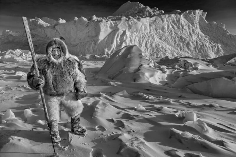 A person in fur clothing stands in a snowy, icy landscape, holding a long stick. Large snow-covered rock formations or icebergs rise in the background under a cloudy sky. The scene is in black and white.