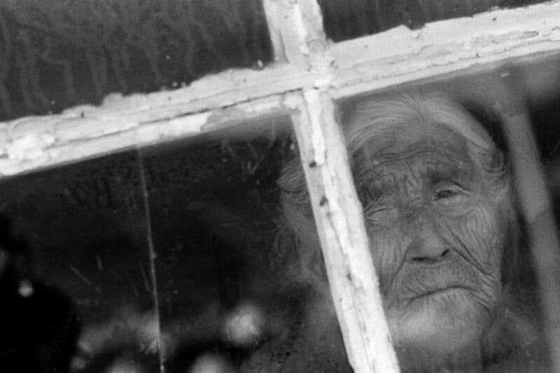 Black and white photo of an elderly person with long hair and a deeply lined face, looking out through a window with a weathered frame and foggy glass.