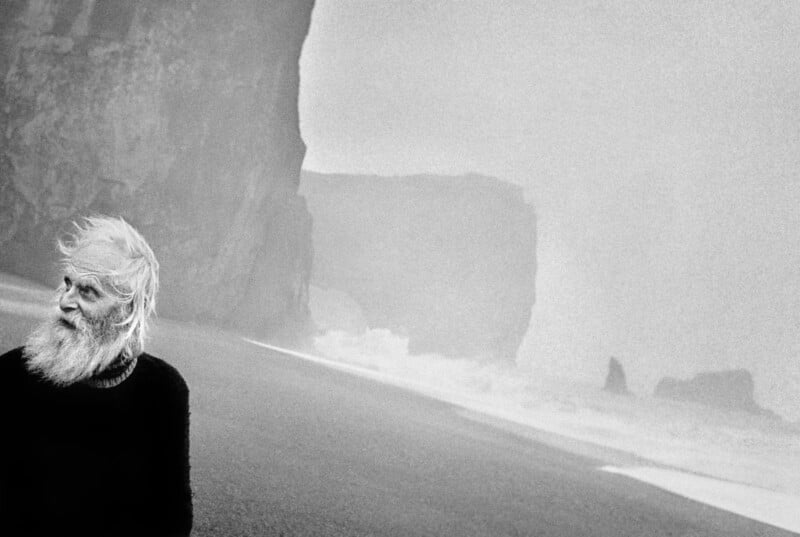An elderly man with long white hair and beard stands on a foggy beach, looking left. Large cliffs and rough waves are in the background, creating a moody, atmospheric scene.