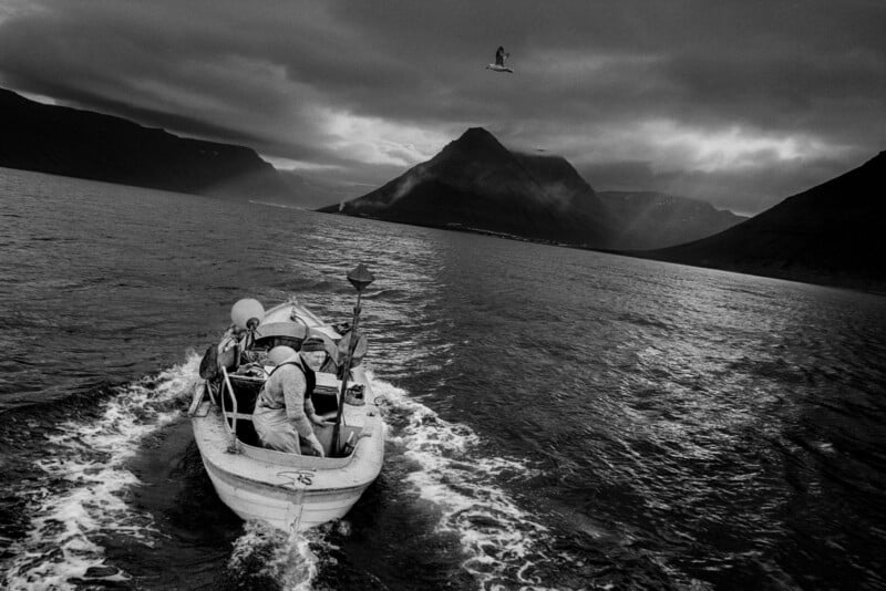 A small fishing boat with several people moves through dark, choppy water toward distant mountains under a cloudy sky. A bird flies above, and the scene is dramatic and moody.