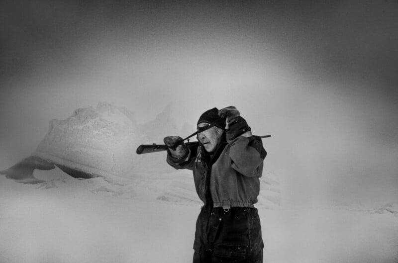 An older person in winter clothing stands in a snowy landscape, looking through a small telescope, with snow-covered mountains and a cloudy sky in the background.
