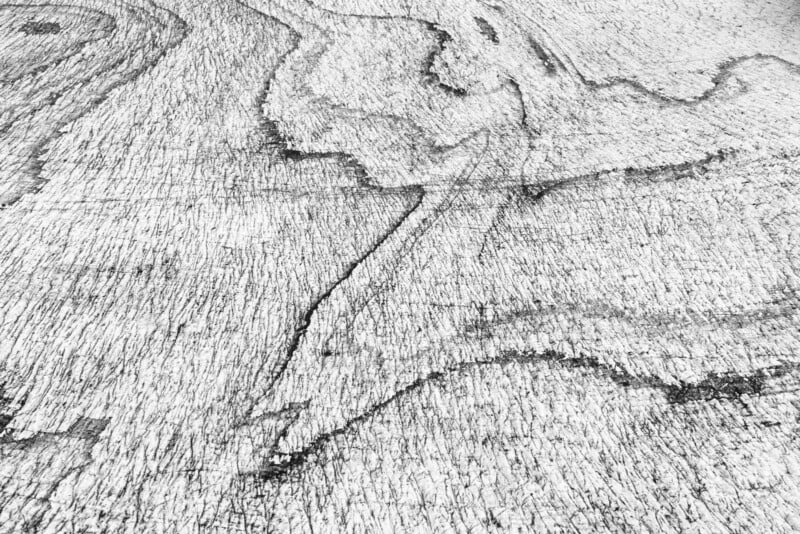 Black and white close-up of a wooden surface with visible grain patterns, lines, and textured marks running across it, creating an abstract and organic appearance.