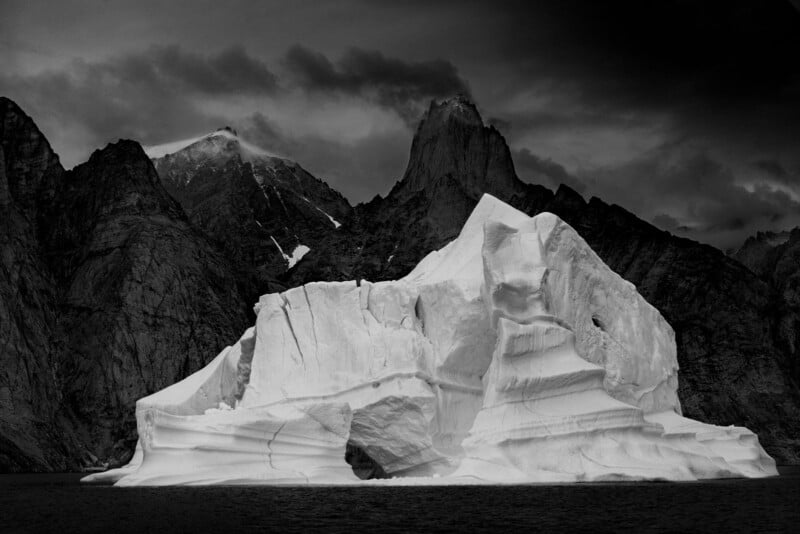A large, jagged iceberg floats in dark water, with steep, rocky mountains and dramatic, cloudy skies in the background. The black and white image emphasizes the stark contrast and textures.