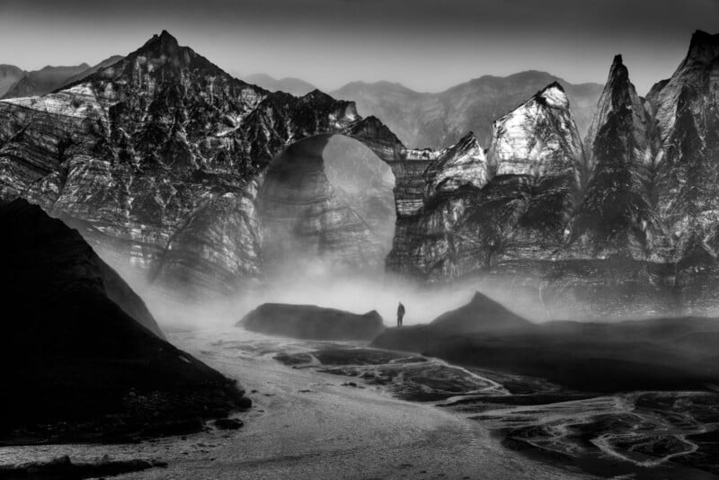A black-and-white image of a dramatic, rugged mountain landscape with a large natural stone arch. Mist swirls at the base, where a lone figure stands on rocky terrain, emphasizing the scale and mystery of the scene.