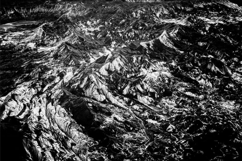 A dramatic black-and-white aerial view of rugged, jagged mountains with sharp peaks and deep valleys, highlighted by contrasting light and shadows.