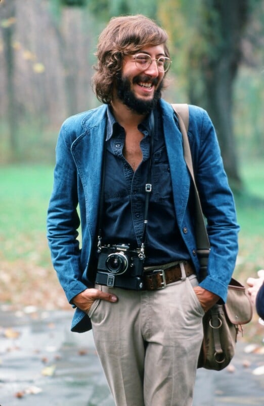 A smiling man with glasses and a beard stands outdoors, wearing a blue jacket, navy shirt, and beige pants. He has a vintage camera around his neck and a brown shoulder bag. The background is blurred greenery.