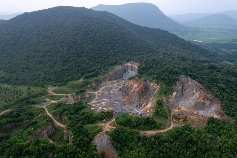 Vista aérea de una gran cantera rodeada de densos bosques y colinas verdes, con paredes rocosas expuestas y caminos de tierra que atraviesan el paisaje. En el interior de la cantera se pueden observar zonas de maquinaria y excavación.