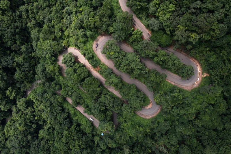 Visto desde un punto de vista elevado, un sinuoso camino de montaña serpentea a través de un denso bosque verde. Se pueden observar varios vehículos circulando por la curva.