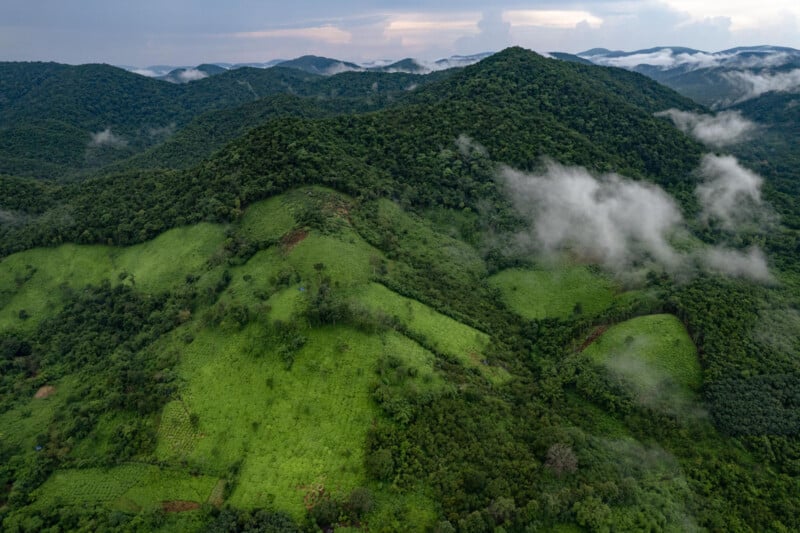Vista aérea de exuberantes montañas verdes, parches de tierras de cultivo, niebla y nubes bajas con colinas distantes y cielo nublado en el fondo.