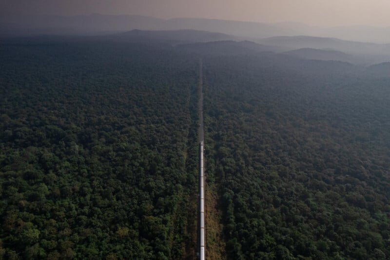 Una vía de ferrocarril recta atraviesa un denso bosque verde y se extiende hacia montañas envueltas en niebla en la distancia bajo un cielo brumoso.