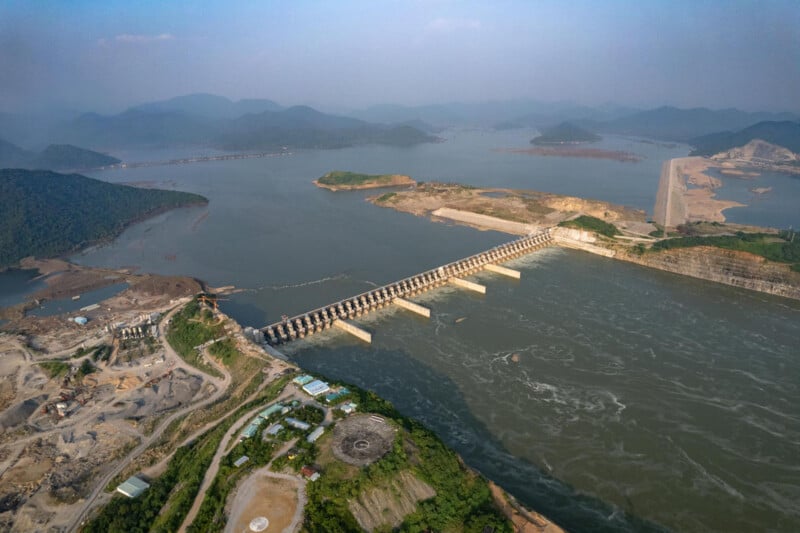 Vista aérea de una presa en un río ancho, rodeada de colinas verdes, sitios de construcción y edificios. Bajo un cielo despejado, las montañas a lo lejos son claramente visibles.