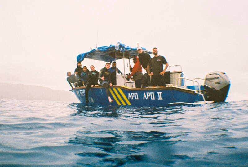 A group of people in wetsuits stand and sit on a small blue boat named "APO APO II" on the ocean, with misty hills visible in the background under a cloudy sky.