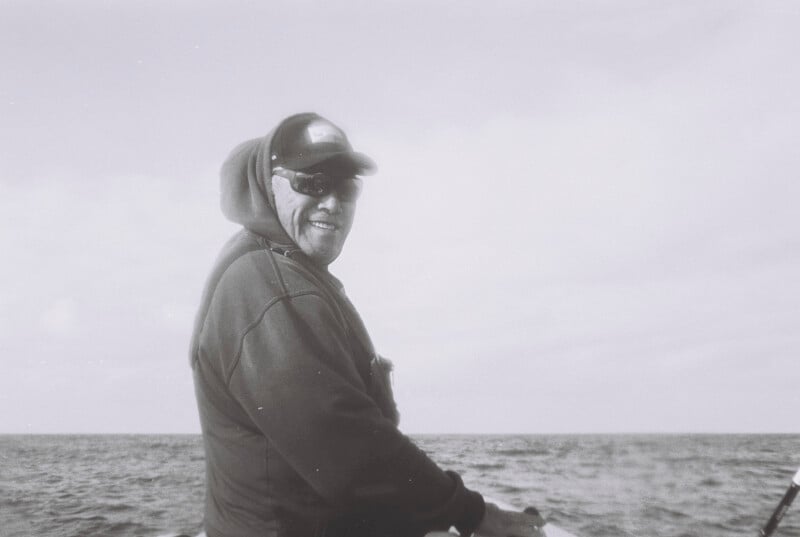 A man wearing sunglasses, a cap, and a hooded sweatshirt stands near the ocean, smiling as he faces the camera. The photo is in black and white, with cloudy skies and the sea in the background.