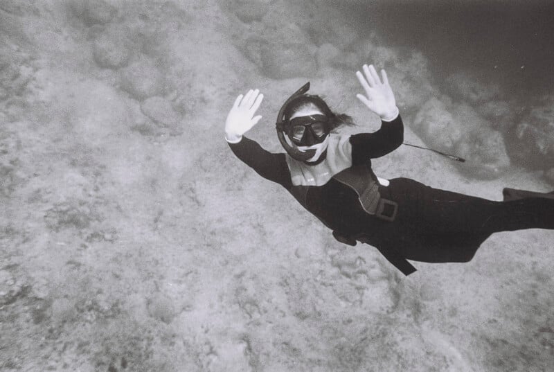 A person wearing a wetsuit, snorkel, and mask swims underwater over a rocky seabed, raising both hands and looking toward the camera. The image is in black and white.