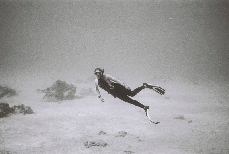 A person wearing a wetsuit, snorkel, mask, and fins swims underwater near the ocean floor, surrounded by rocks and sand in a calm, clear environment.