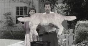 A man in a vintage outfit holds a large golden retriever horizontally over a grill, while a smiling woman stands beside him in front of a house. The image is in black and white.