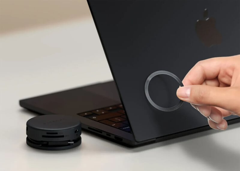 Una mano aplica un círculo de adhesivo en la parte posterior de una computadora portátil Apple negra. Hay un accesorio negro redondo sobre una mesa cercana.