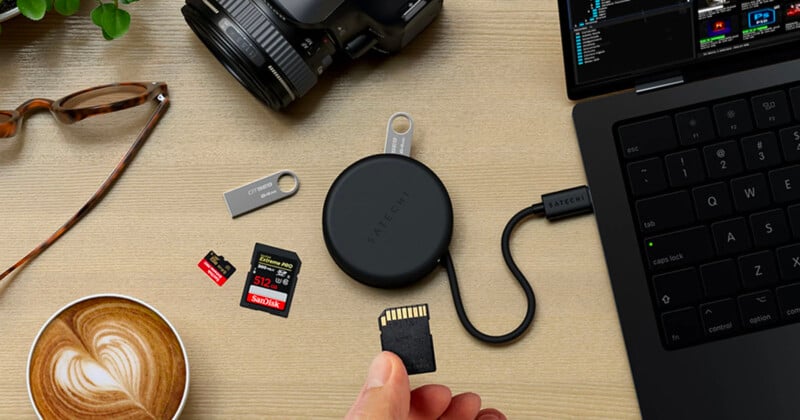 Hay una tarjeta SD encima del lector de tarjetas sobre una mesa con una computadora portátil, una cámara, una unidad USB, una tarjeta de memoria, vasos, una planta y un café con leche con una obra de arte en forma de corazón.