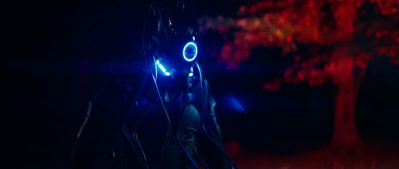 Una figura que viste un traje brillante futurista con luces azules y cables se encuentra en la oscuridad, mirando hacia adelante. En el fondo borroso se puede ver un árbol con hojas iluminadas en rojo.