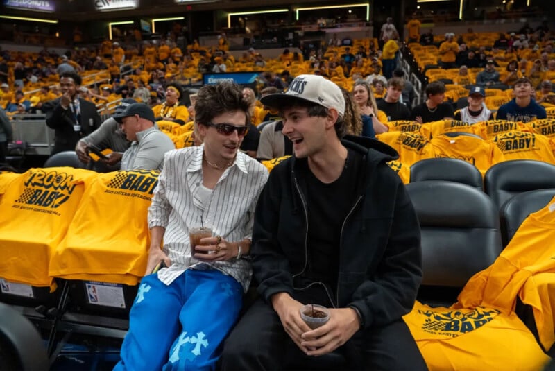 Dos jóvenes estaban sentados al margen de un partido de baloncesto, sonriendo y sosteniendo bebidas. Los asientos detrás de ellos estaban cubiertos con camisetas amarillas y otros fanáticos se sentaban detrás de ellos.