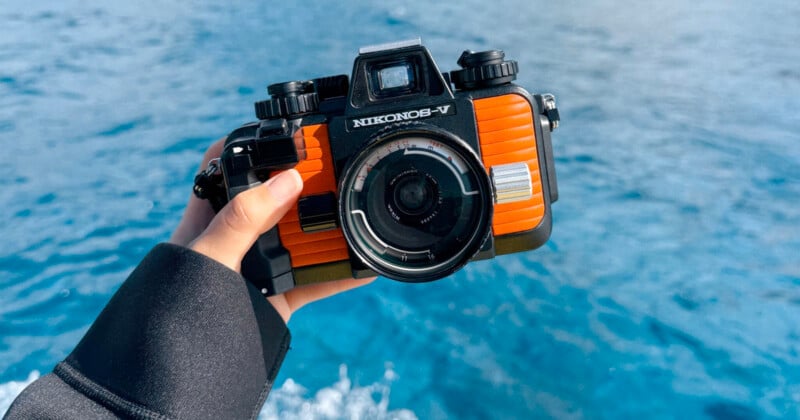 A hand holding a black and orange Nikonos-V underwater camera with a blurred blue water background.