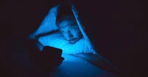 A person lies in bed under a blanket at night, staring at a smartphone that emits a blue glow, illuminating their face in the dark room.
