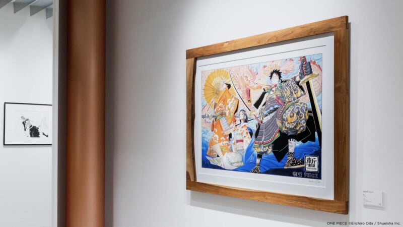 Una colorida obra de arte enmarcada cuelga de una pared blanca y presenta figuras con trajes elaborados. Otra imagen monocromática más pequeña es parcialmente visible a la izquierda. El escenario parece ser una galería o corredor moderno.