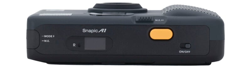 Top view of a dark gray Snapic A1 camera showing its yellow shutter button, mode and M.E. switches, a small display screen, and the on/off switch. The lens is visible in the center.