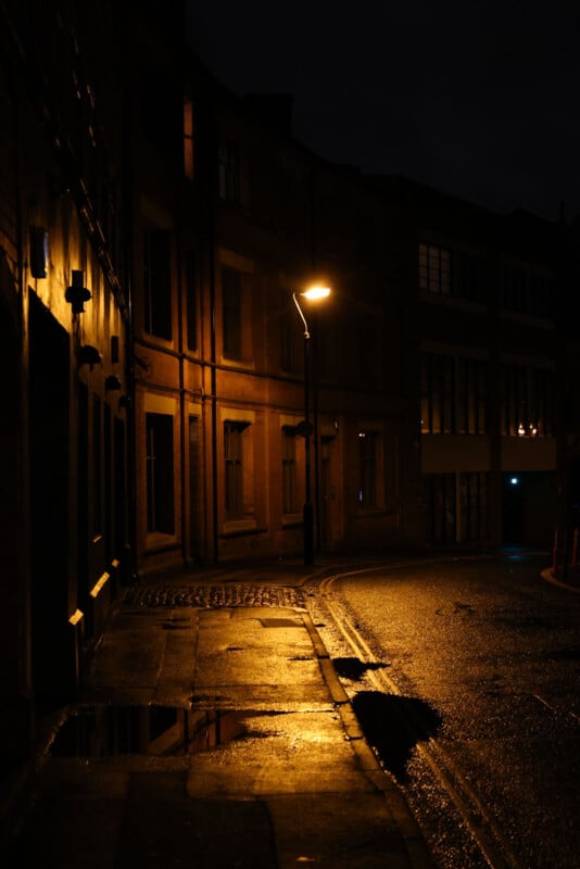 Por la noche, las luces de la calle son tenues, la carretera está mojada y hay muchos edificios de gran altura. Una farola proyecta luces y sombras sobre la acera y la carretera.