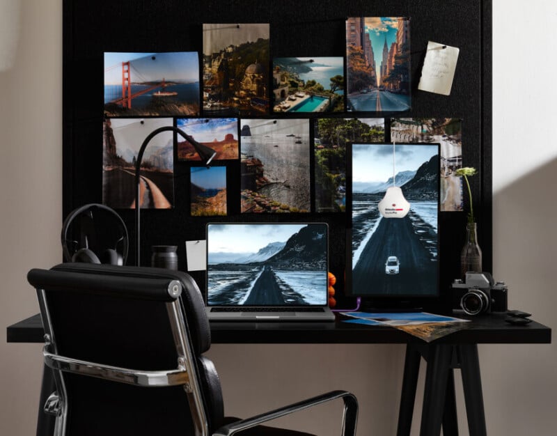 A black desk with a laptop, vertical monitor, camera, headphones, and notebooks sits against a wall covered in travel photos, creating a cozy workspace with a black office chair in front.