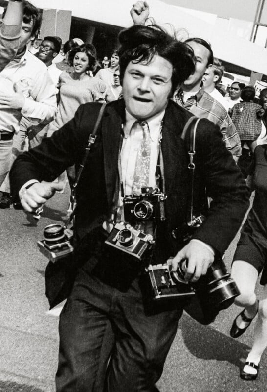A man runs forward with multiple cameras around his neck, looking determined. A crowd of people follows behind him, some appearing excited or energetic. The image is black and white.