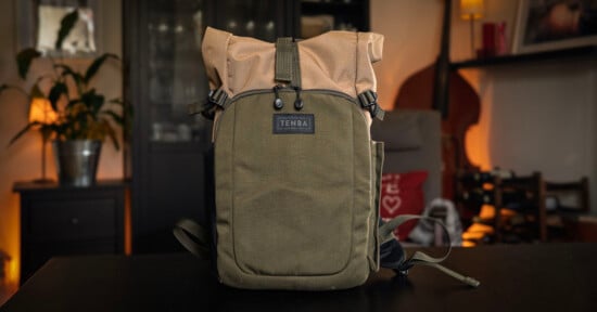 A green and tan Tenba backpack sits on a dark table in a cozy, softly lit room with plants, a bass, and shelves in the background.