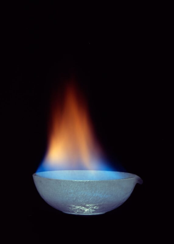 A white ceramic bowl with a blue and orange flame rising from it, set against a completely black background.