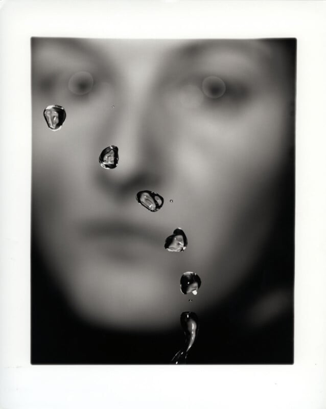 A black and white blurry portrait of a person’s face, with several water droplets on the surface. Each droplet sharply reflects and magnifies a portion of the face underneath, creating a surreal, fragmented effect.