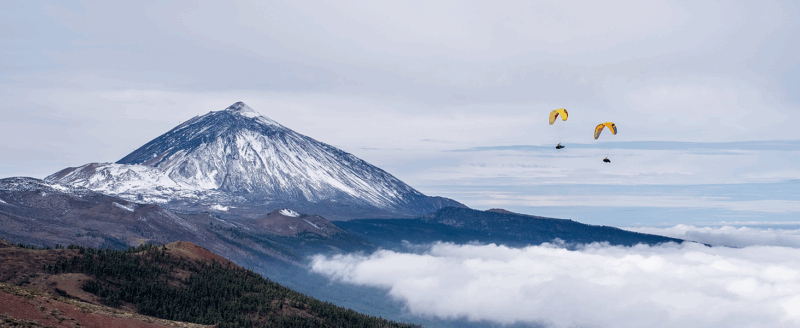 Dos personas se deslizan con paracaídas amarillos sobre las nubes bajo un cielo nublado con montañas nevadas y colinas boscosas al fondo.