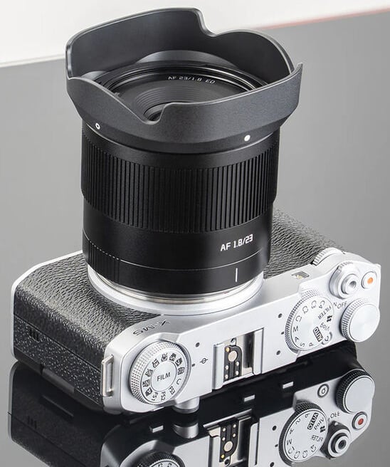 Una cámara sin espejo plateada y negra equipada con una gran lente gran angular descansa sobre una superficie lisa, mostrando claramente los diales de control, los botones y parte del parasol.