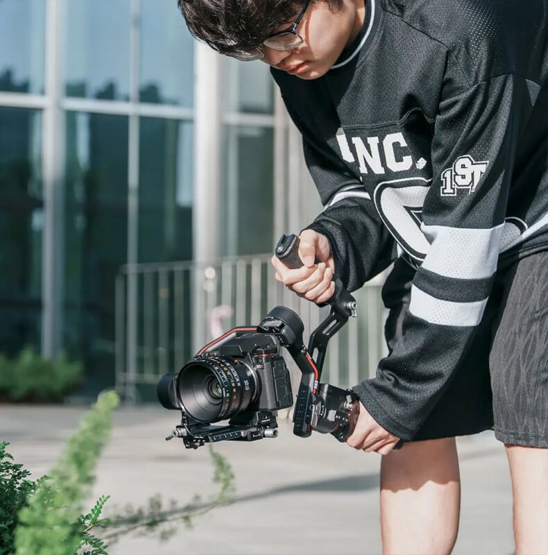 Un hombre con gafas y una sudadera negra opera una cámara profesional montada en un estabilizador para filmar plantas al aire libre cerca de un edificio con ventanas de vidrio.
