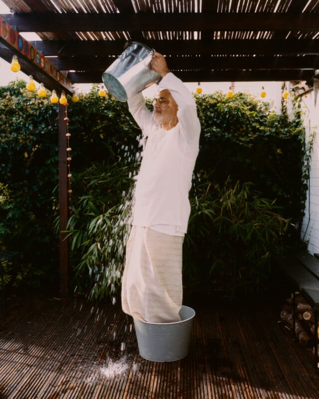 An older man in white clothing stands in a metal bucket outdoors, pouring water over himself from another bucket, with greenery and a wooden pergola in the background.