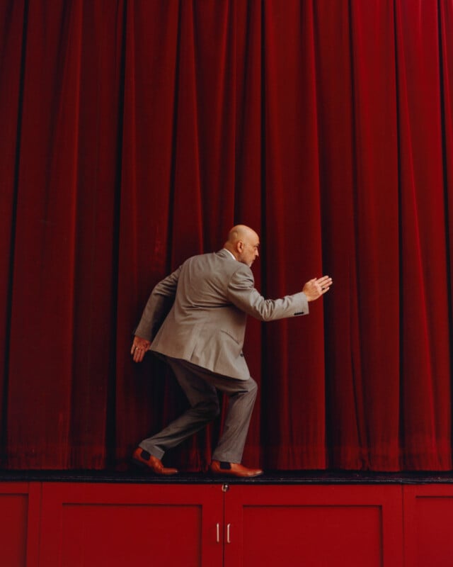 A man in a gray suit and brown shoes steps out from behind large red stage curtains, with one hand extended forward, against matching red walls and floor.